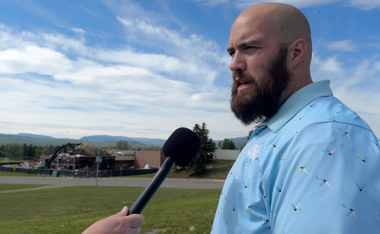 default-1359 a bald, bearded man in a light blue patterned shirt gives an outdoor interview with mountains and a school building being demolished behind him