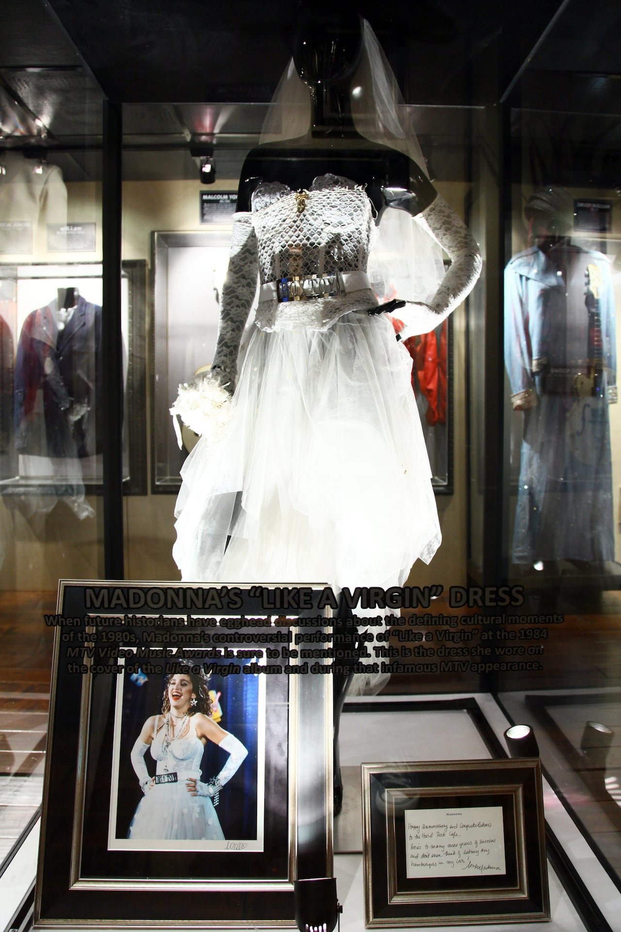 default-1434 A wedding dress and a belt reading "Boy Toy" displayed in a case on a mannequin. A photo of a woman wearing the dress and a plaque sit in front of it.