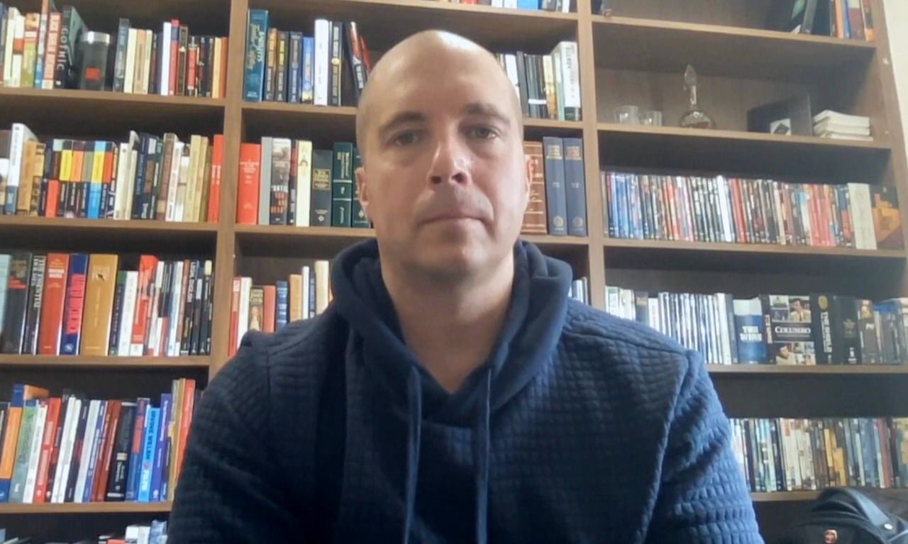 A bald man in a blue hooded sweater sitting in front of a bookshelf.