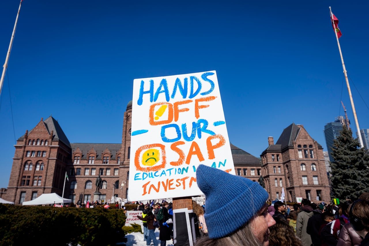 In front of Queen's Park legislative building in downtown Toronto on a blue-sky winter day, a large crowd rallies. A prominent sign in the foreground reads Hands Off Our OSAP