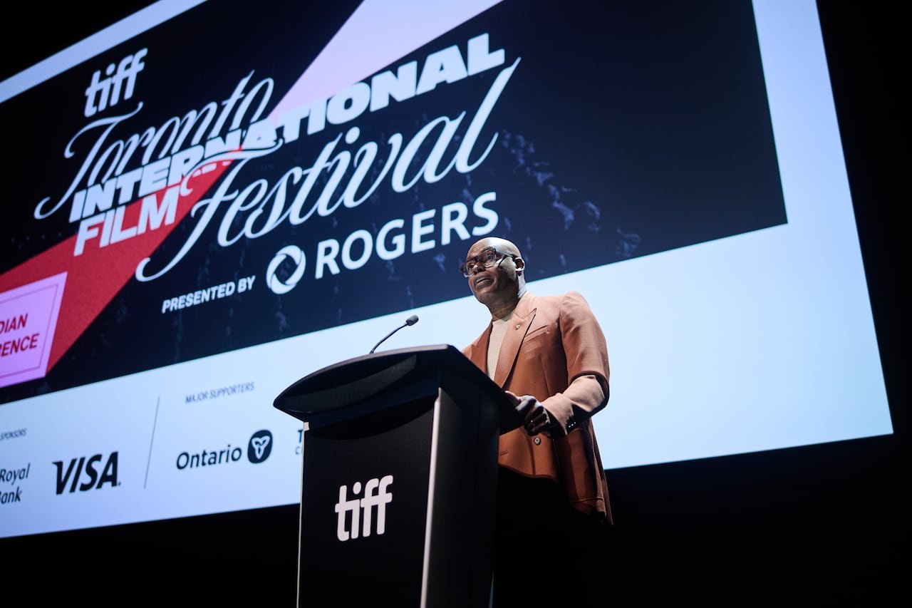 A bald man wearing glasses speaks at a podium in front of a large screen advertising the Toronto International Film Festival.