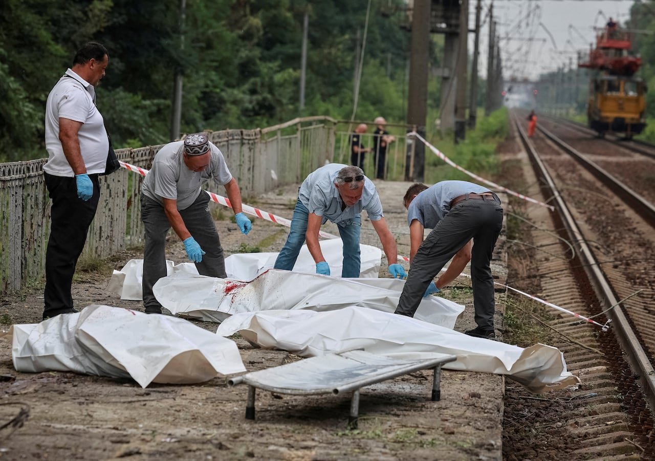 Four men are show moving around bodies inside white body bags next to a train track.