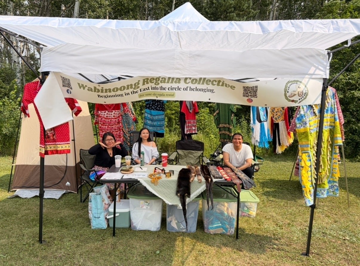 Three individuals in a vendor tent with regalia under the 'Wabinoong Regalia Collective' banner
