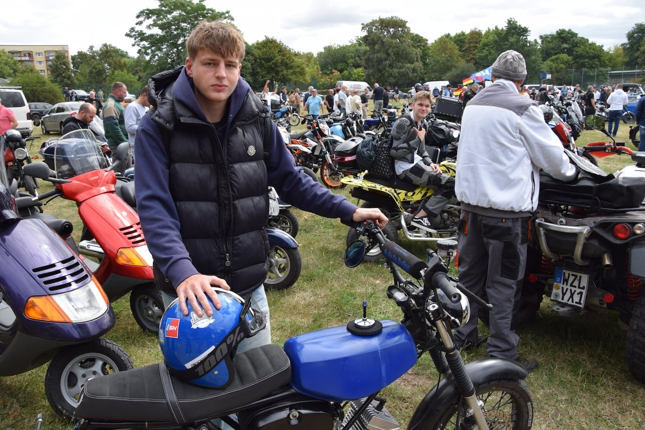 A teenage boy wearing a purple sweater and a black vest stands behind a blue and black motorcycle.