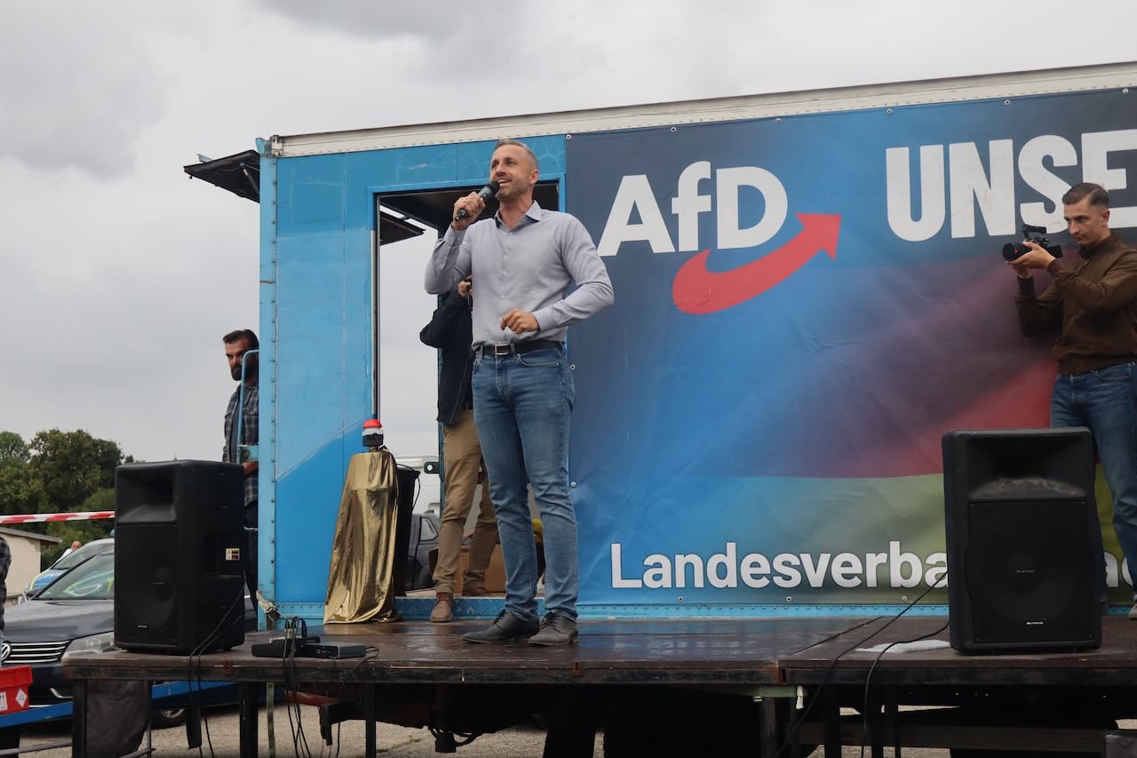 A man wearing jeans and a button-up blouse stands on a stage and speaks into a microphone.