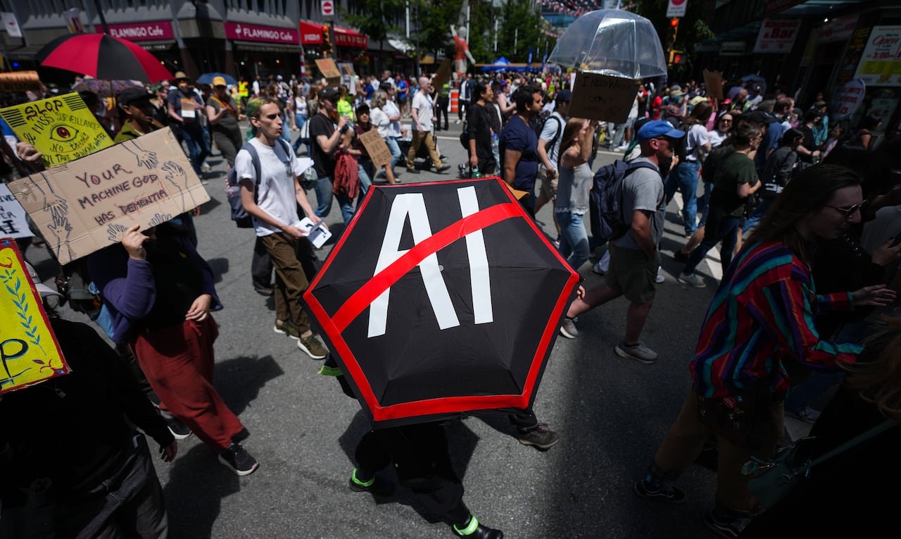 A large group of people are outside protesting. At the centre is an umbrella that has "AI" with a big slash over it.