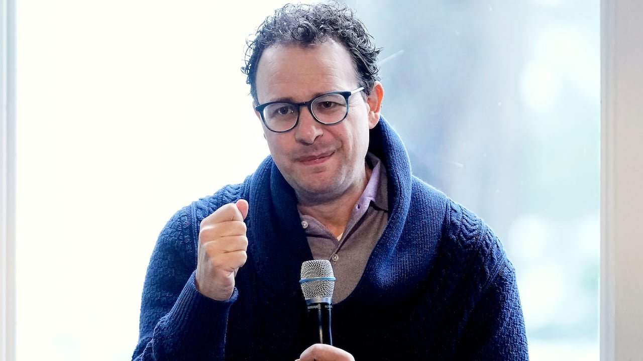 A man holds a microphone with his other fist held up, making a closed-mouth half-smile.