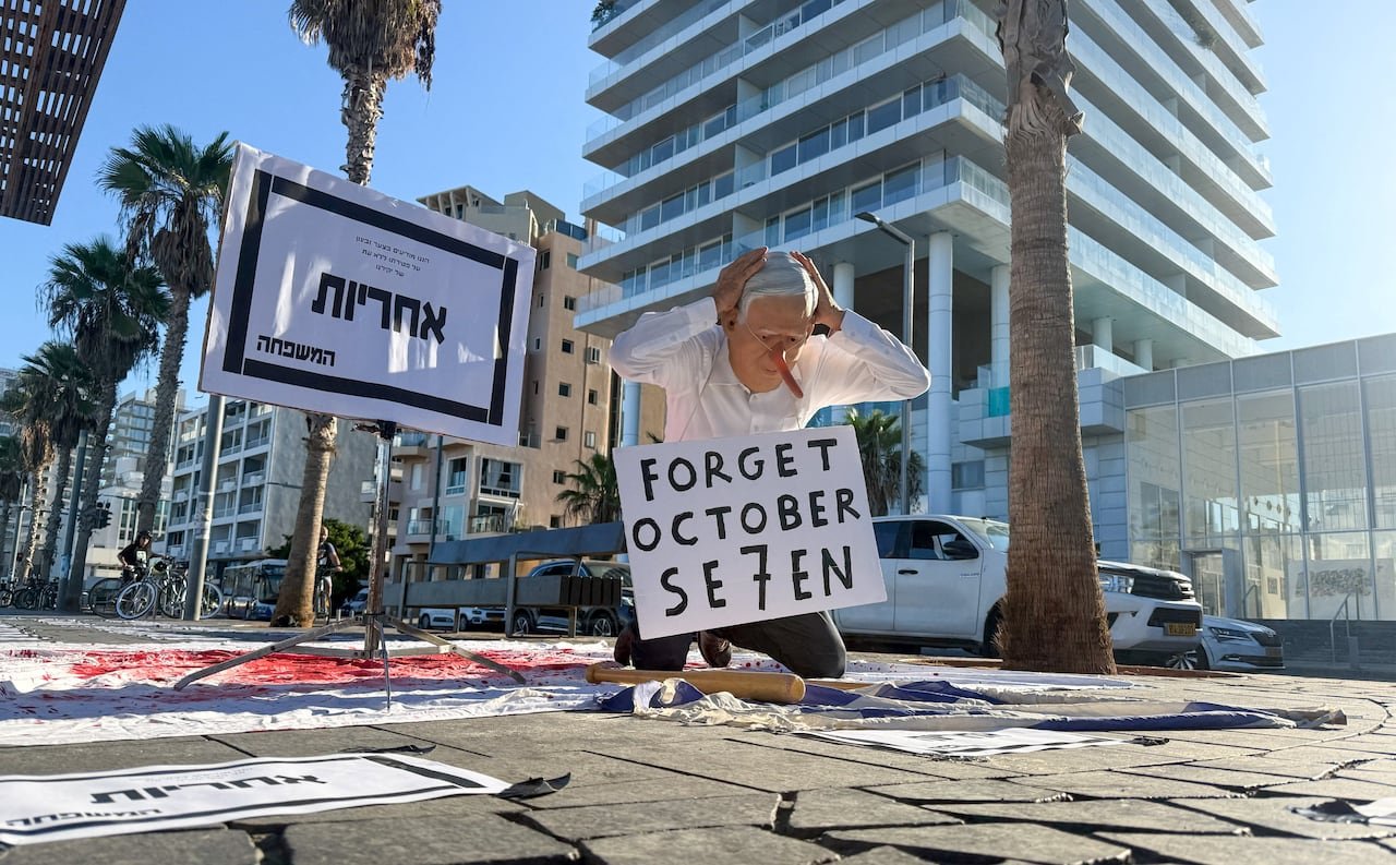 A protester in a full-face mask kneeling on a city street with a sign that reads 'Forget October Seven' around his neck.