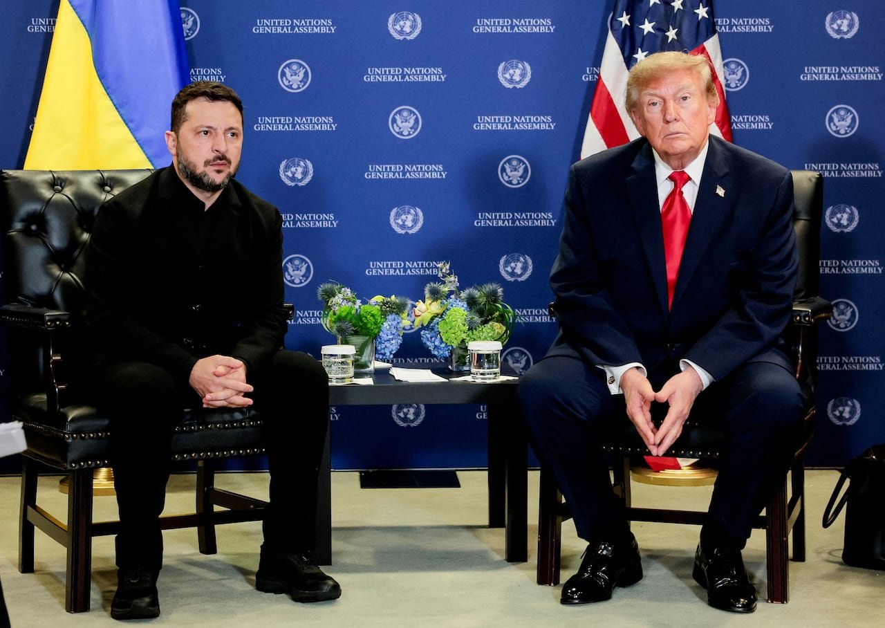 default-1889 Two men sit, with their hands folded on their laps, in front of the flags for Ukraine and the United States and a blue wall with the words "United Nations General Assembly" on it.