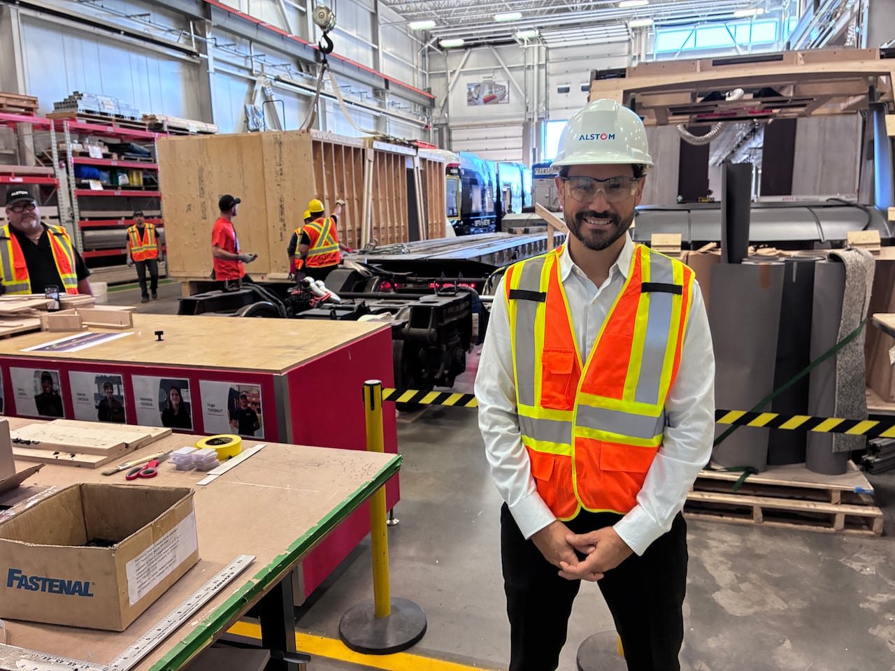 A man in an orange safety vest, goggles, and a hard hat stands on a factory floor.