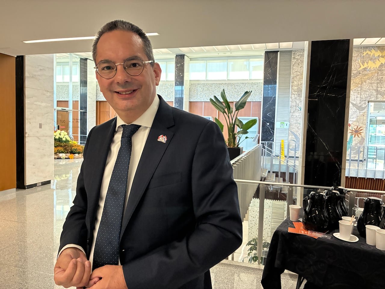 A man in a dark colored suit stands and smiles at the camera.