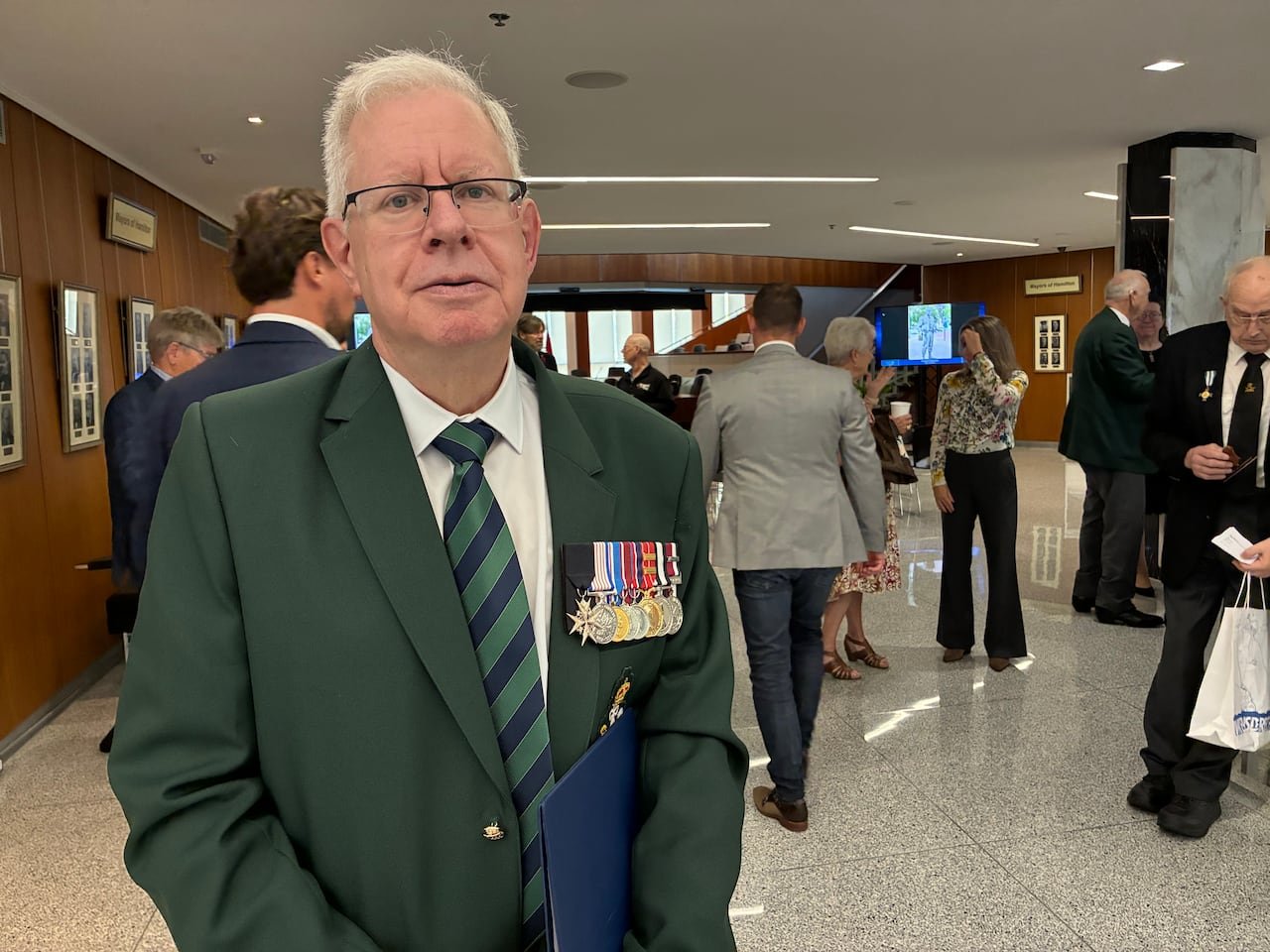 A man wearing a green suit with military medals stands looking at the camera.