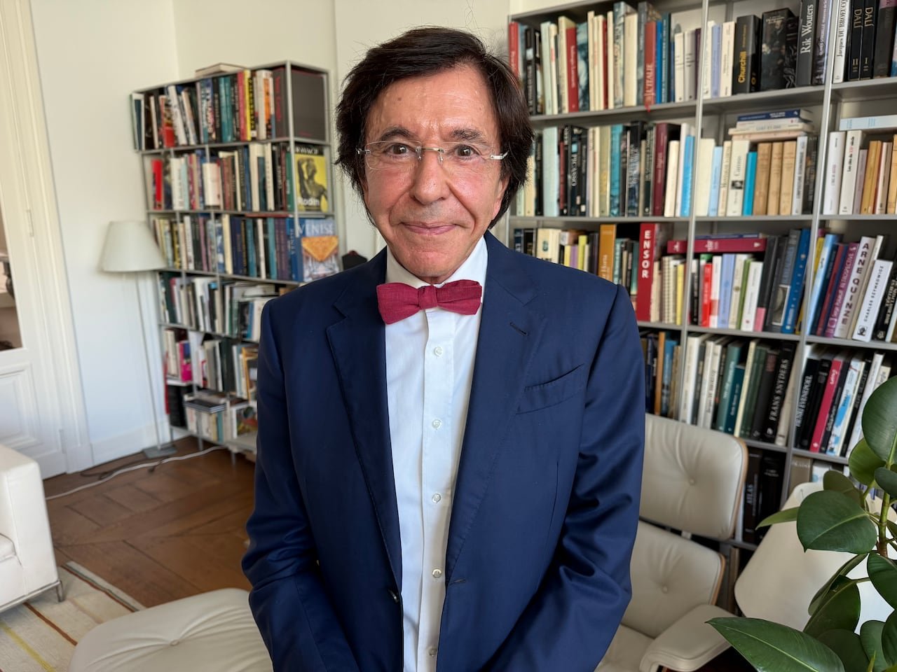 A man with glasses, dark hair, red bow-tie, and blazer standing in front of a book shelf.