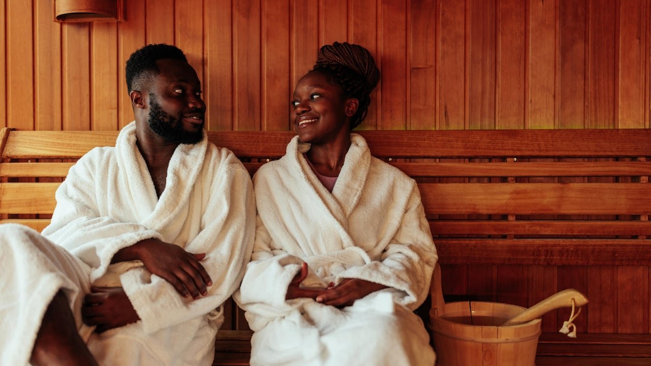 A couple in bath robes, sitting in a wood-paneled sauna, smiling at each other.