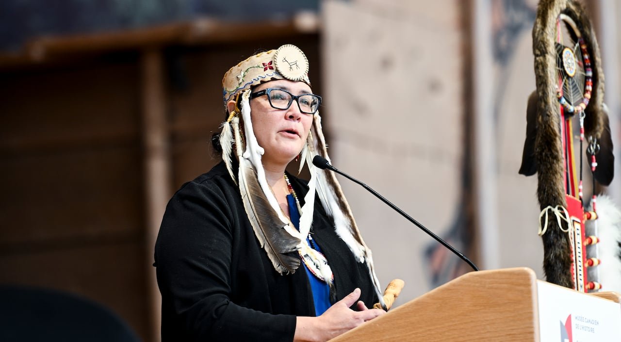 A woman speaks into a mic at a podium outdoors with an eagle staff beside her.