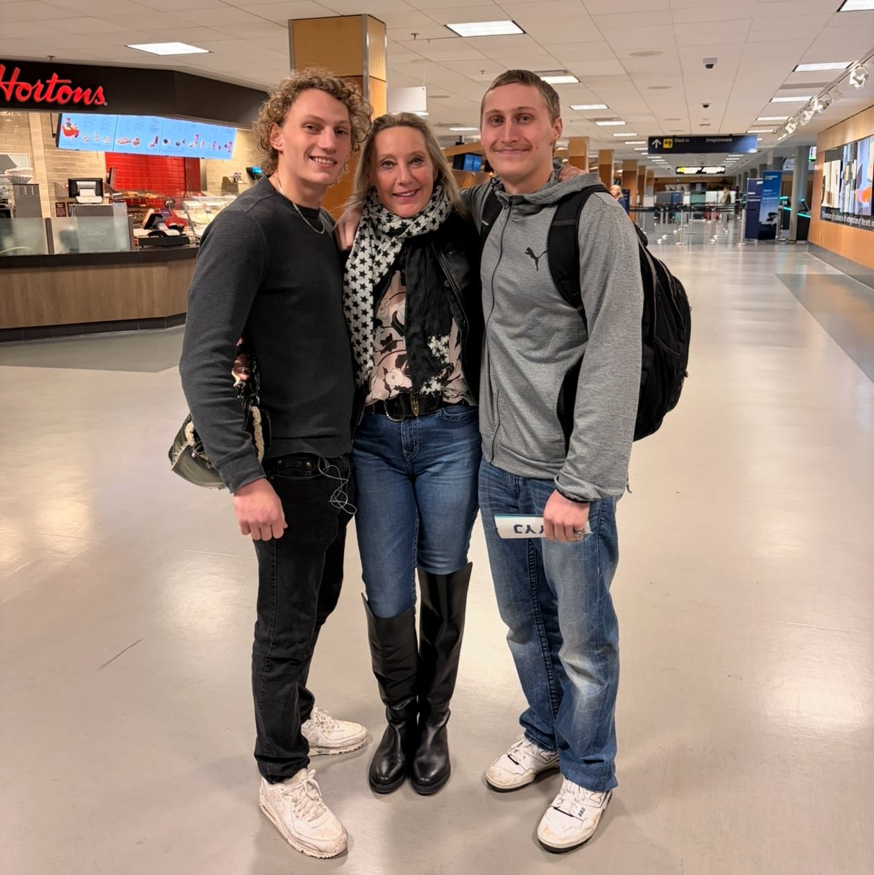 A woman with long blonde hair poses with her arms around two smiling young men in front of a Tim Hortons inside an airport.
