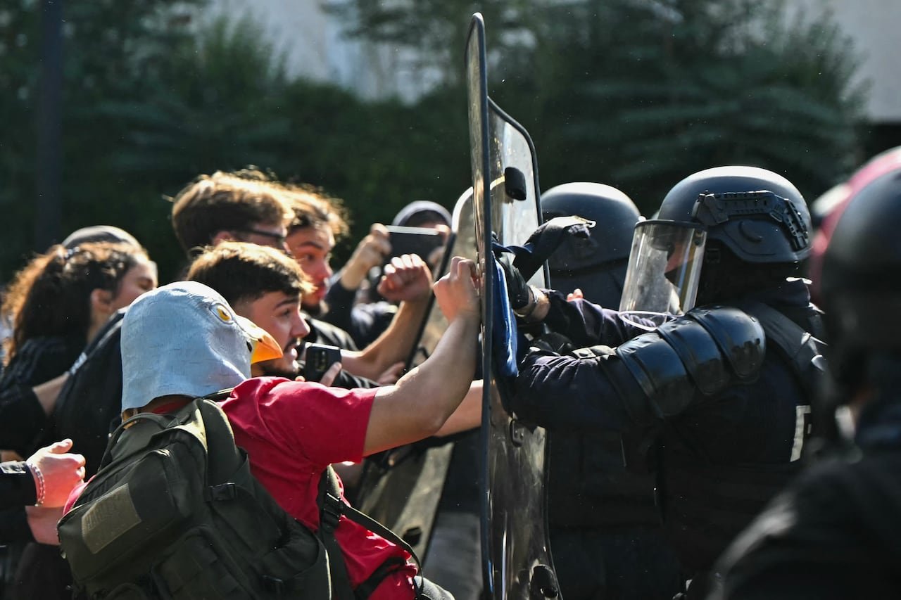Young men and women push against riot shields held aloft by riot police.