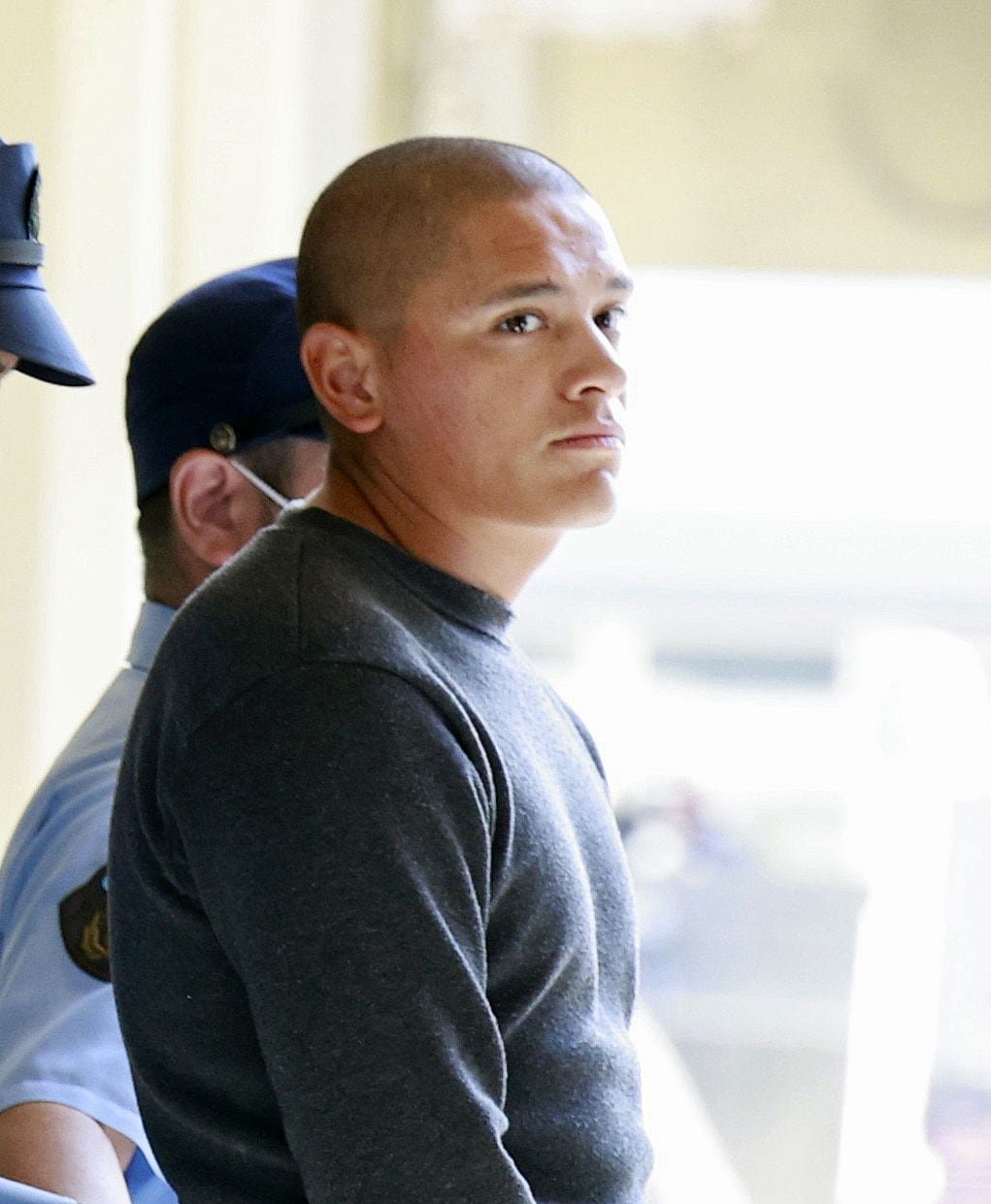 A muscular man with a shaved head, wearing a gray sweater, flanked by two unformed police officers, looks to one side with a serious expression.